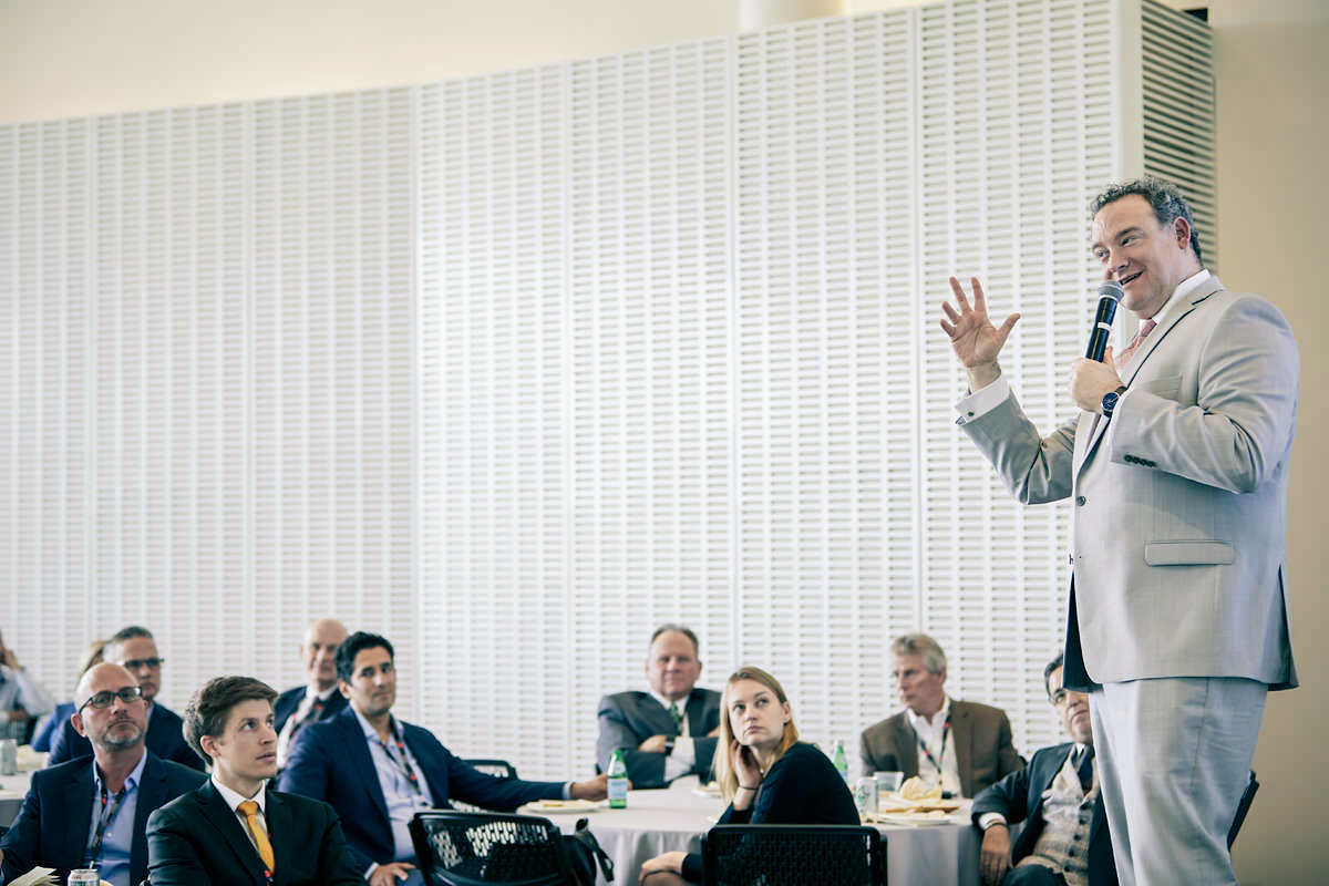 People seated at tables look at a man standing and talking into a microphone.