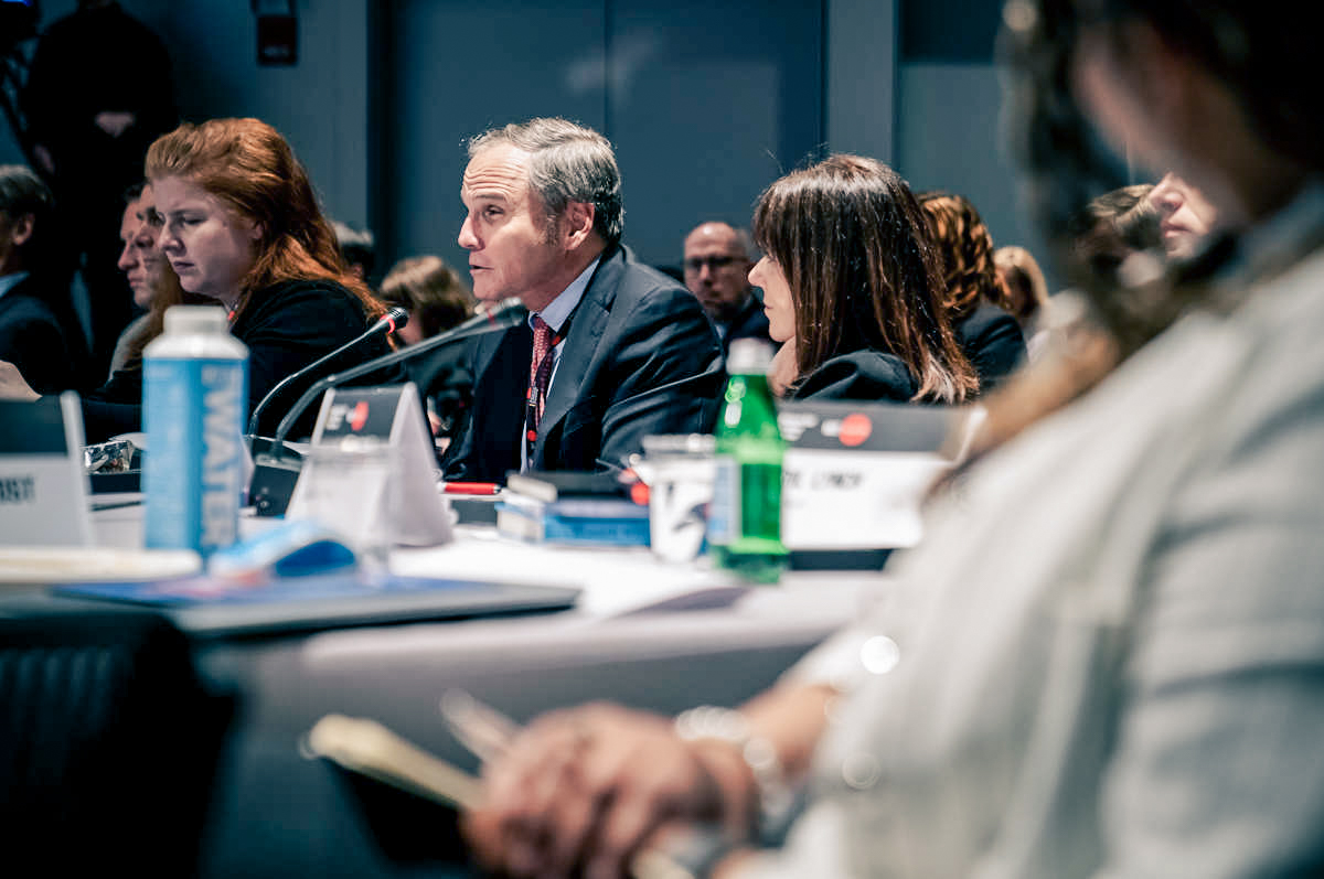 Older man sits at a panel table in front of a microphone with women sitting on either side, and people sitting behind.