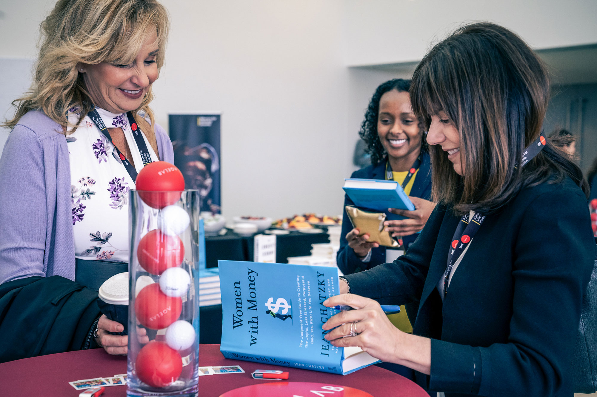 Woman signs a book at a table for another woman.