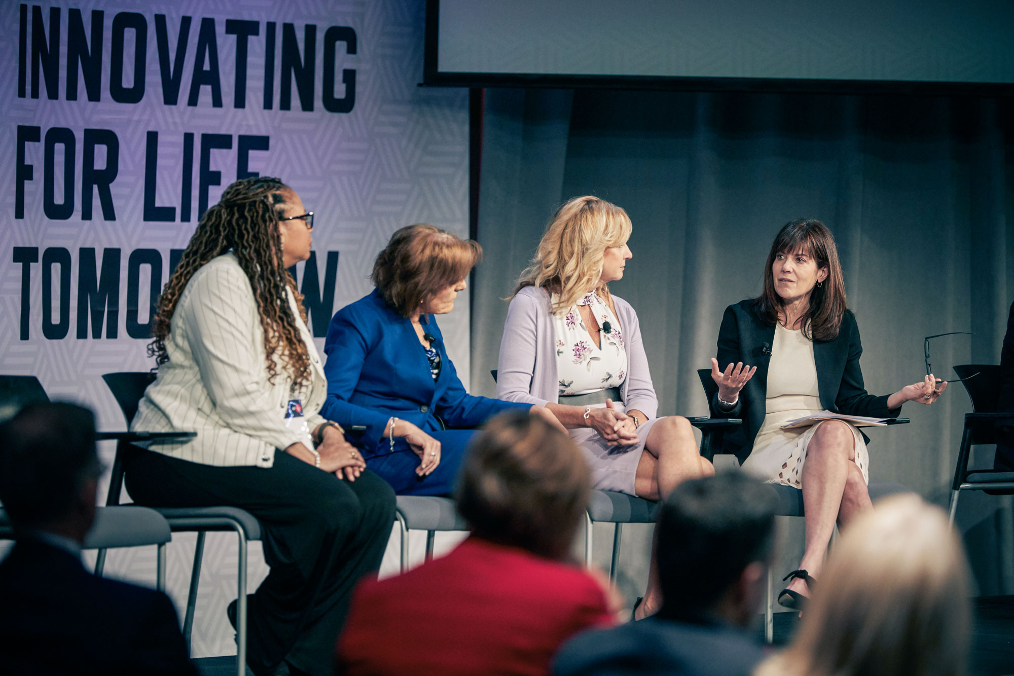 4 women sit in chairs on a stage and one is speaking while the others look toward her