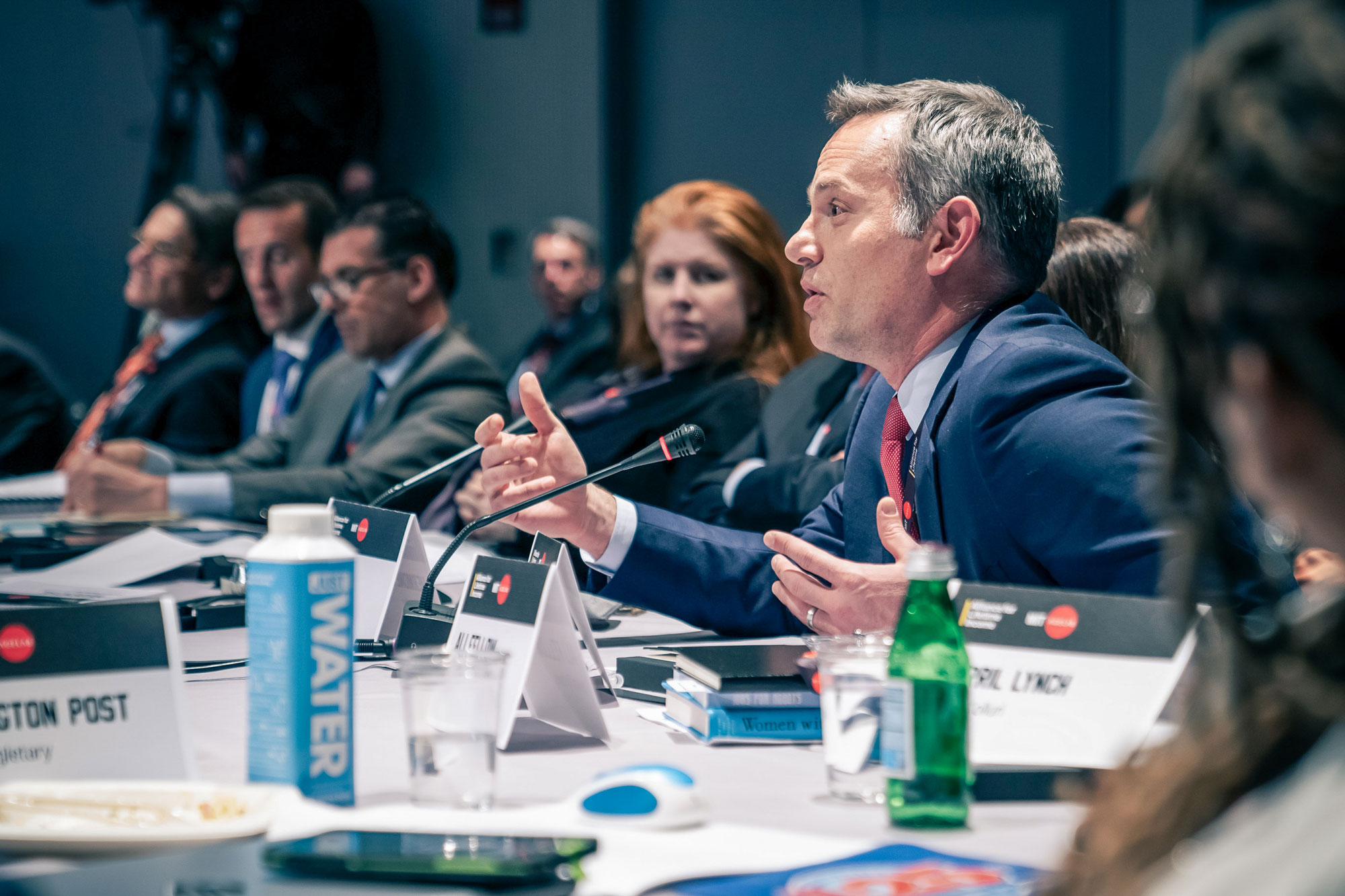 Man talks into a microphone while sitting at a panel table with others.